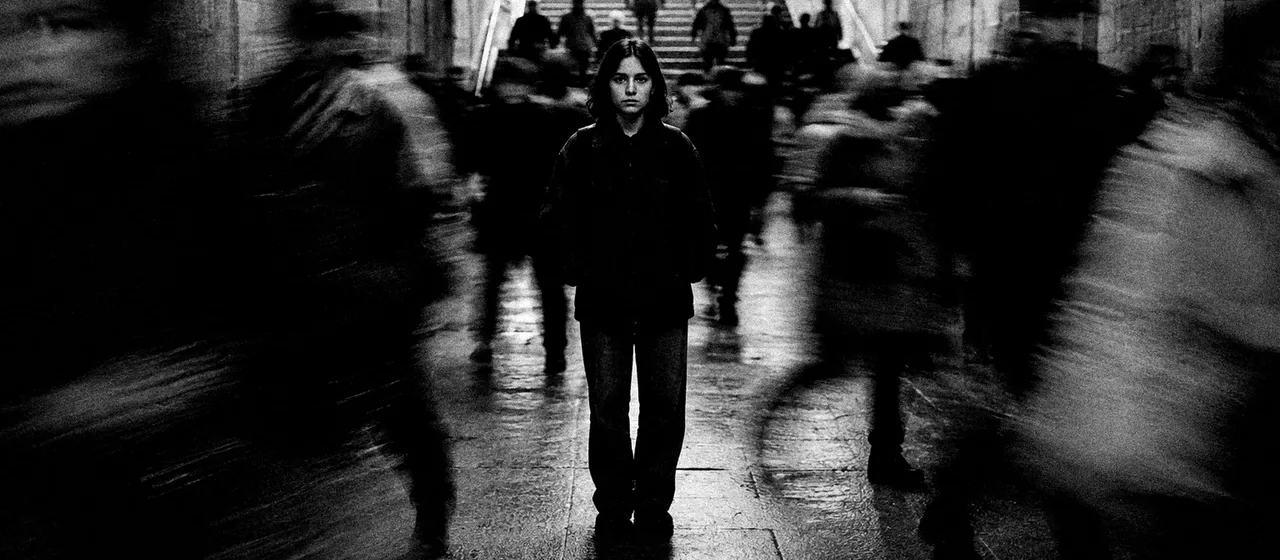 En blanco y negro, una chica quieta y nítida en medio de la multitud que pasa movida a su lado, en un paso subterráneo.