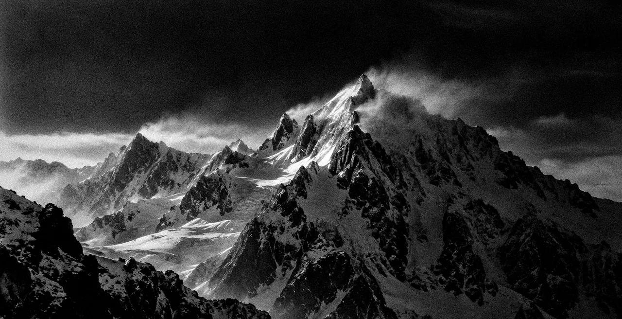 Cumbres nevadas en blanco y negro bajo un cielo oscuro, el viento levanta la nieve de la cima más alta.