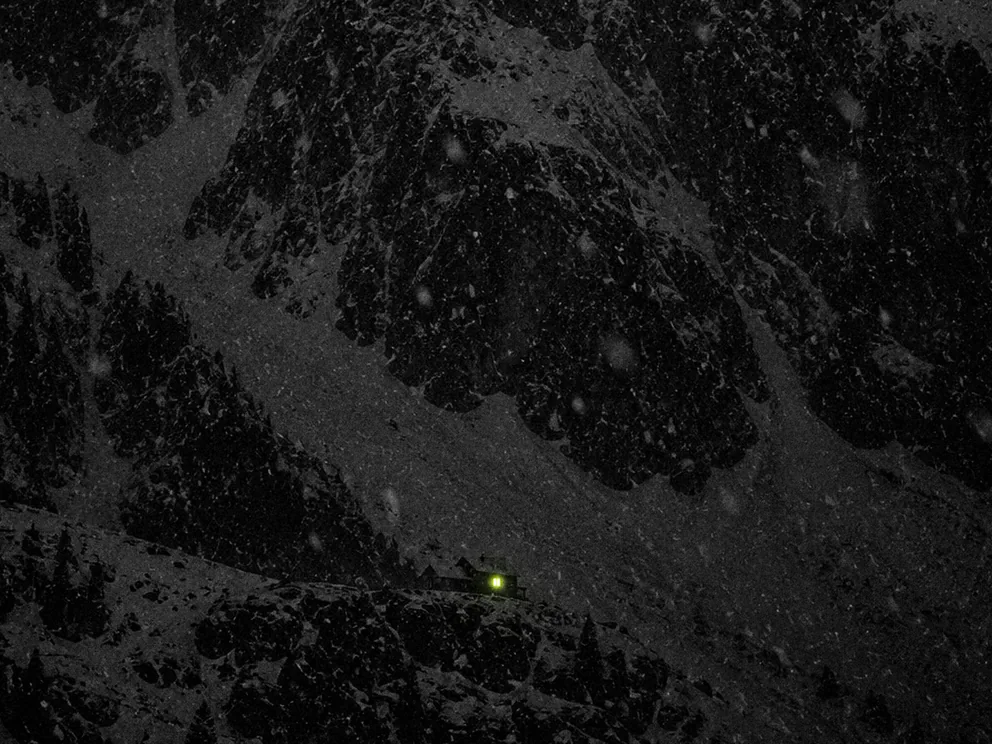 De noche, bajo la nieve, una montaña enorme y a sus pies un refugio con una sola ventana encendida.