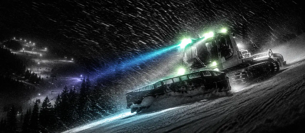 Una máquina pisanieves prepara la pista de noche, con los faros encendidos bajo la nieve que cae.