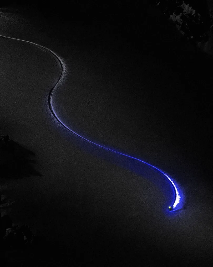 Una sola huella de esquí baja en curva por una ladera oscura, con una estela azul.