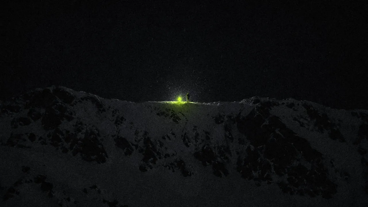 De noche, en la cresta de una montaña, una persona diminuta junto a un fuego lima: la única luz encendida.
