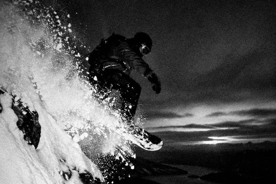 Un snowboarder salta desde una cresta de noche, en una nube de nieve polvo, con el lago y la última luz debajo. Foto en blanco y negro.