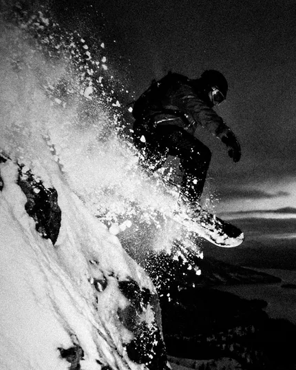 Uno snowboarder in curva alza un muro di neve, in bianco e nero.