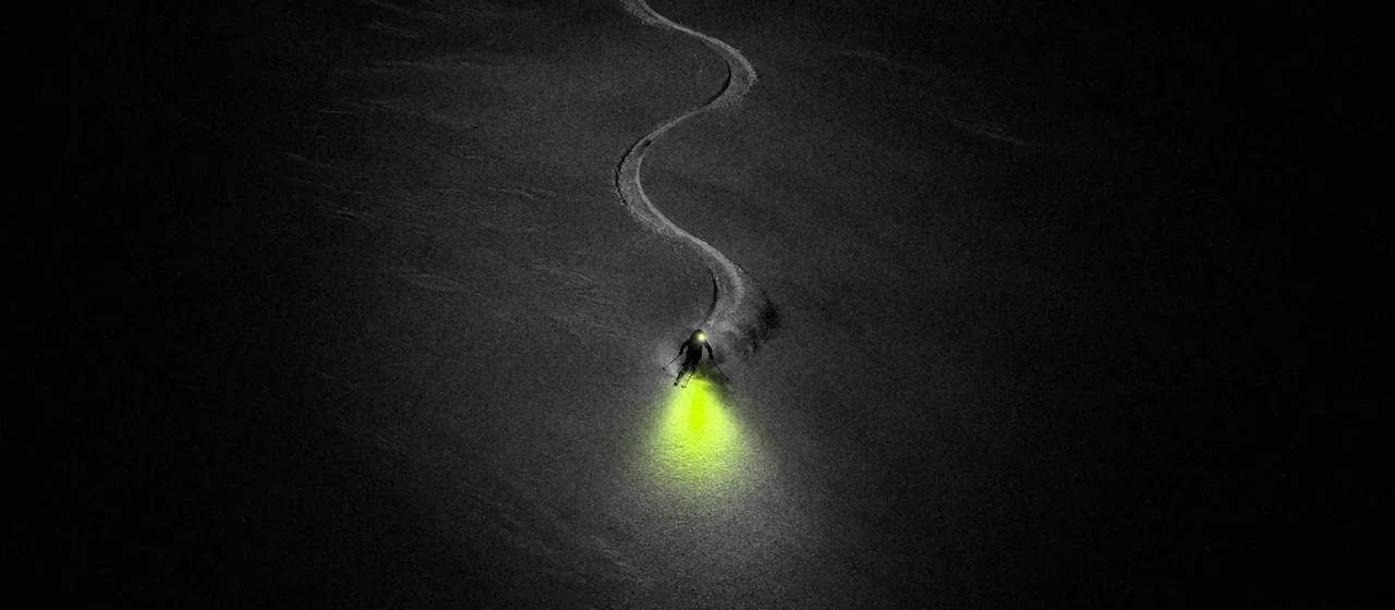 De noche, en una ladera de nieve virgen, un esquiador baja con el frontal encendido: detrás de él un solo surco, delante un charco de luz lima.